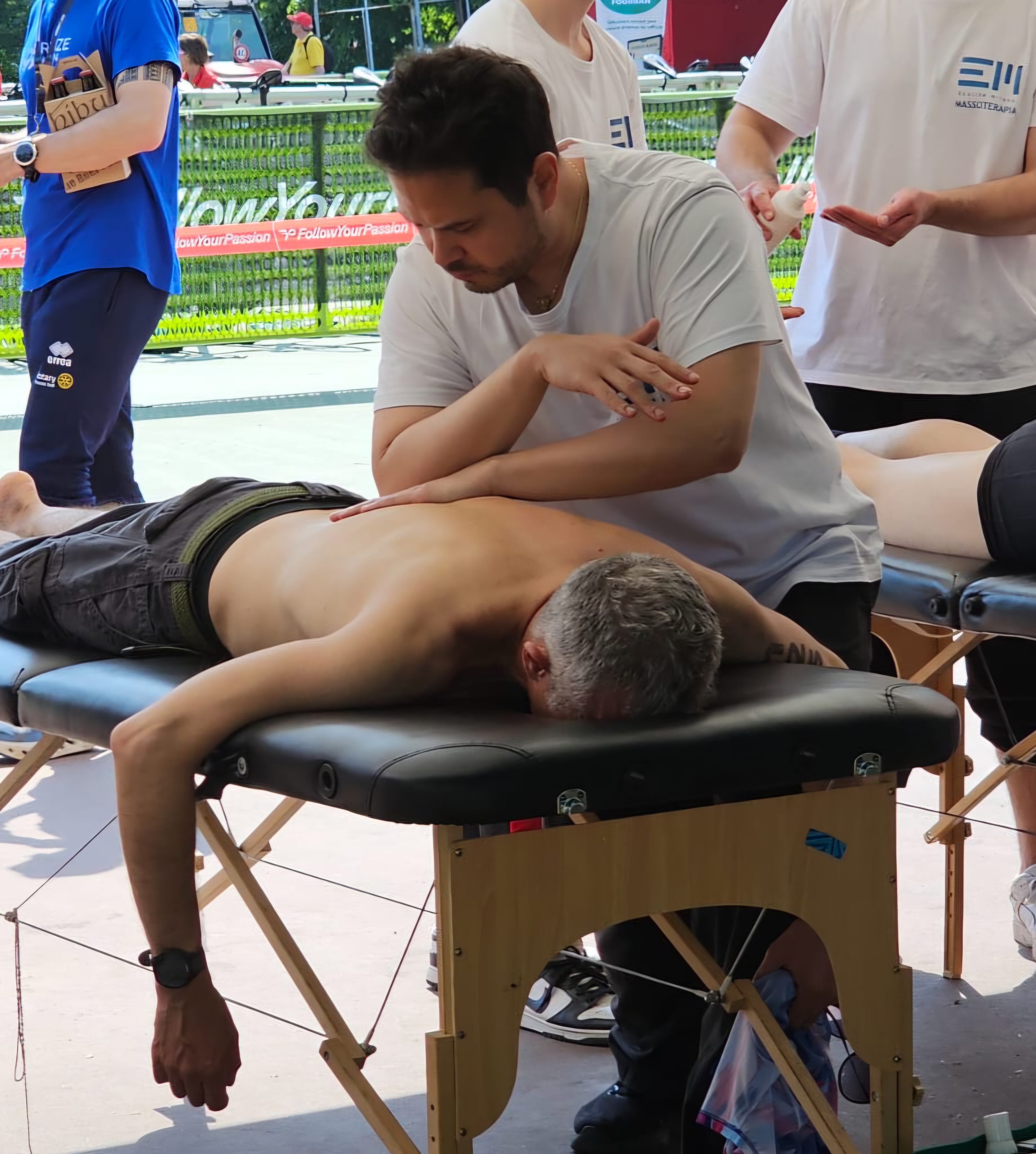 Un operatore sta eseguendo un massaggio a un uomo sdraiato su un lettino. Sullo sfondo, si vedono altre persone coinvolte in attività simili.
